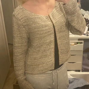 Beige stickad kofta - Säljer en beige stickad kofta med guldig detaljer vid öppningen. Koftan har långärmad design och en rak, lite boxig passform. Perfekt att slänga över en topp för en chill och snygg look. Aldrig använd💕