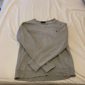 Grå sweatshirt från Polo Ralph Lauren - Säljer en stilren grå sweatshirt från Polo Ralph Lauren i bomull. Tröjan har rund hals, långa ärmar och den klassiska broderade loggan på bröstet. Perfekt för en chill och clean look.