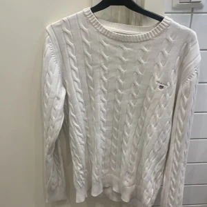 Vit kabelstickad tröja från Gant - Kabelstickad vit tröja från Gant med rund halsringning och diskret logga på bröstet. Tröjan har långa ärmar och ribbade muddar vid ärmslut och nederkant. Perfekt för lager-på-lager och klassisk preppy stil.