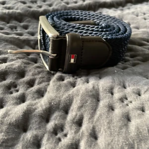 Mörkblått Tommy Hilfiger bälte - Snyggt mörkblått bälte från Tommy Hilfiger med vävd struktur och detaljer i svart skinn. Spännet är i silverfärgad metall och bältet har en liten logotyp vid lädret. Perfekt accessoar för att lyfta din outfit.