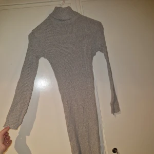 Grå ribbad stickad långklänning - Mysig grå långklänning i ribbstickat material med polokrage och långa ärmar. Klänningen har en figurnära passform och är perfekt för kyligare dagar. Enkel och stilren design som är lätt att matcha med olika accessoarer.