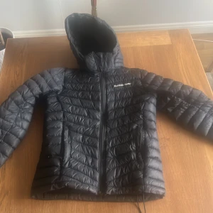 Svart dunjacka med huva från Peak Performance - Säljer en svart dunjacka från Peak Performance med snygg quiltad design och huva. Jackan har dragkedja framtill och två sidofickor. Perfekt för kalla dagar och har en modern, sportig look som funkar till de flesta stilar.