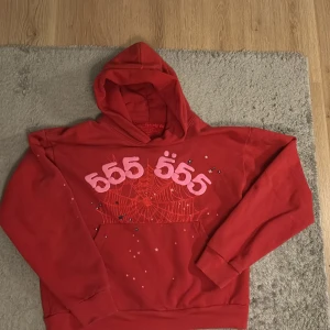 Röd SP5DER hoodie med 555-tryck - Röd hoodie från SP5DER med stor huva och fet 555-print i rosa framtill. Trycket har en spindelnätsdesign och små stjärnor, plus coolt SP5DER-tryck på huvan. Mjuk insida och klassisk känguruficka. Perfekt statement-plagg för streetwear.