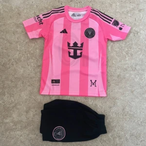 Inter Miami Messi #10 rosa fotbollströja - Säljer en Inter Miami fotbollströja från Adidas i rosa med svarta detaljer och ränder. Tröjan har korta ärmar, klubbmärke och sponsortryck, samt Messi och nummer 10 på ryggen. Materialet är lätt och andas, perfekt för match eller träning.