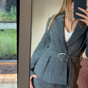 Grå kavaj med bälte från NA-KD - Snygg grå kavaj från NA-KD med markerad midja och stort silverfärgat spänne. Materialet är mjukt och känns som ullblandning. Perfekt till kostymbyxor för en cool look.