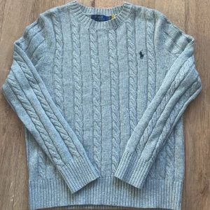 Grå kabelstickad tröja Polo Ralph Lauren - Kabelstickad grå tröja från Polo Ralph Lauren med klassisk rund halsringning och broderad logga på bröstet. Tröjan är långärmad och har ribbade muddar vid ärmslut och nederkant. Perfekt för lager-på-lager under kyliga dagar.