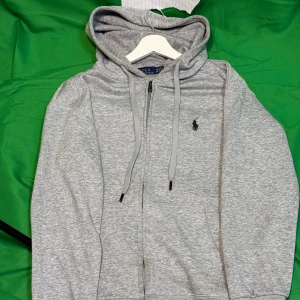 Grå hoodie från Polo Ralph Lauren - Snygg grå hoodie från Polo Ralph Lauren med dragkedja framtill, huva med snörning och klassisk broderad logga på bröstet. Mjuk bomullsblandning och känguruficka gör den både chill och stilren. Perfekt till jeans eller joggers.Liknande modell som originalet!