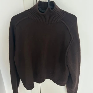Brun stickad polotröja från H&M - Mörkbrun stickad polotröja från H&M med hög krage och ribbade detaljer vid hals och nederkant. Tröjan har lång ärm och en något croppad passform, perfekt för kyligare dagar. Den är kort i modellen men jag skulle inte säga att det är en magtröja.