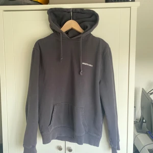 Mörkgrå hoodie från Axel Arigato - Säljer en mörkgrå hoodie från Axel Arigato med vit textprint både fram och bak. Tröjan har huva med snörning, känguruficka och är tillverkad i 100% ekologisk bomull.
