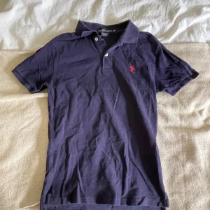 Mörkblå pikétröja från U.S. Polo Assn. - Säljer en klassisk mörkblå pikétröja från U.S. Polo Assn. i bomull. Tröjan har korta ärmar, krage och knäppning framtill samt röd broderad logga på bröstet. Perfekt för en stilren och sportig look.