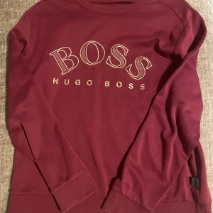 Vinröd sweatshirt från Hugo Boss - Säljer en vinröd sweatshirt från Hugo Boss med stort BOSS-tryck i guld på bröstet. Tröjan har rund hals, långa ärmar och ribbade muddar. Perfekt för dig som vill ha en stilren och trendig look med premiumkänsla. 