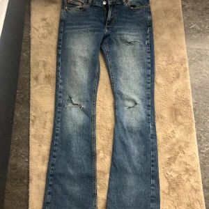 Blå bootcut jeans från Junkyard S - Snygga blå bootcut jeans från Junkyard i storlek S. Jeansen har slitna detaljer vid knäna och råa, fransiga benslut för en trendig look. Klassisk femficksmodell med orange sömmar och normal passform. Perfekta för dig som gillar en avslappnad stil.