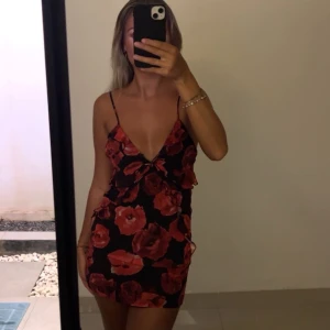 Svart kortklänning med röda blommor - Säljer en svart kortklänning med röda blommor och djup v-ringning. Klänningen har tunna axelband och en figurnära passform som framhäver siluetten. Perfekt för dig som vill sticka ut med ett snyggt blommigt mönster och en feminin look.