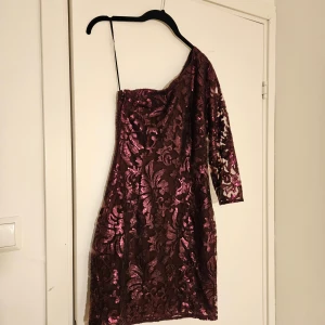 Glittrig vinröd one shoulder klänning - Säljer en vinröd glittrig klänning från H&M i storlek 36. Klänningen har ett snyggt one shoulder-snitt med en lång ärm och är täckt av paljetter i ett blommigt mönster. Bara använd en gång 
