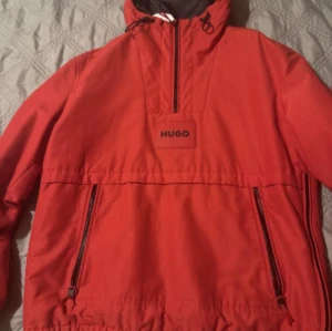 Röd vindjacka från Hugo med huva - Riktigt snygg röd vindjacka från Hugo med svart innerfoder och huva. Jackan har dragkedja framtill, två sidofickor med dragkedja och justerbar snörning vid huvan. Perfekt för dig som vill sticka ut med en clean och sportig look.