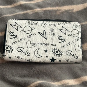 Vit plånbok med doodle-print från FSBN - Cool vit plånbok från FSBN med svart doodle-print, fylld med ögon, hjärtan, stjärnor och texter. Plånboken har dragkedja runtom och flera fack för kort och mynt. Materialet är syntet med struktur och insidan är svart.