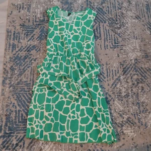 Grön och vit mönstrad klänning - Säljer en kort klänning med grönt och vitt grafiskt mönster som påminner om giraffmönster. Klänningen har ärmlös design, markerad midja och rund halsringning. Perfekt för dig som gillar färgstarka och unika plagg.