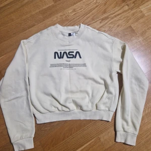 Beige NASA sweatshirt från H&M - Säljer en beige sweatshirt från H&M med stor svart NASA-logga framtill. Tröjan har rund hals, långa ärmar och ribbade muddar vid ärmslut och nederkant. Perfekt för dig som gillar rymd och streetstyle.