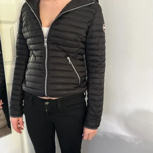 Svart dunjacka från Colmar storlek S - Snygg svart dunjacka från Colmar med quiltad design och silvriga dragkedjor. Jackan har huva, två fickor med dragkedja och Colmar-logga på ärmen. Perfekt för kyliga dagar och ger en clean, sportig look.