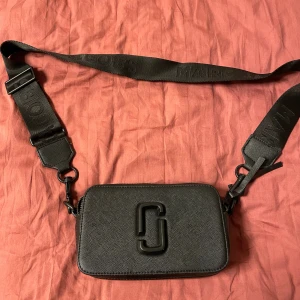 Svart axelväska Marc Jacobs  - Snygg svart axelväska från Marc Jacobs i strukturerat skinn med bred justerbar axelrem med logga. Väskan har dubbla dragkedjor, diskret logotyp framtill och en praktisk ficka på baksidan. Perfekt och tuff.