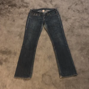 True Religion mörkblå jeans, stl 31w - Mörkblå jeans från True Religion med klassiska kontrastsömmar och snygga bakfickor med lock och knappar. Jeansen har rak passform och är tillverkade i kraftigt denim. Made in USA. Perfekta för dig som gillar street och premiumkänsla. En defekt som visas på bild, men den e fixad så bra det går. Inte säker på längd men gissar på 32-34