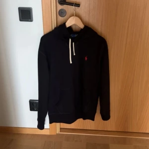 Svart hoodie från Ralph Lauren - Svart hoodie från Ralph Lauren med vit snörning och klassisk röd logga på bröstet. Tröjan har huva, känguruficka och långa ärmar. Perfekt för en chill och stilren look. Tillverkad i mjuk bomull för extra komfort.