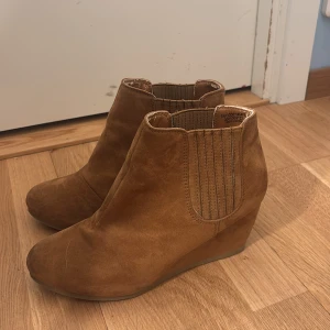 Bruna mocka chelsea boots med kilklack - Snygga bruna chelsea boots i mocka med bred passform och bekväm kilklack. Elastiska paneler på sidorna gör dem enkla att ta på och av. Perfekta för dig som vill ha både stil och komfort i vardagen.