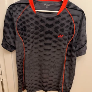 Svart och grå sport-tshirt från Yonex - Sportig t-shirt från Yonex i svart och grått med coolt mönster och röda detaljer längs sömmarna. Tillverkad i 100% polyester, perfekt för träning. Kortärmad modell med rund hals och snygg kontrastkrage i rött. Storlek M.