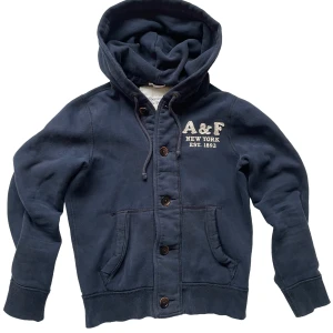 Mörkblå hoodie från Abercrombie & Fitch - Mörkblå hoodie från Abercrombie & Fitch med broderad A&F-logga och text på bröstet. Tröjan har knäppning med stora knappar, huva med snörning och två fickor framtill. Skön bomullskvalitet och ribbade muddar vid ärmslut och nederkant.