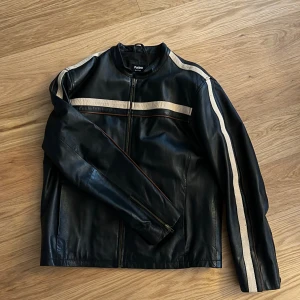 Svart skinnjacka med vita ränder Patino - Svart skinnjacka från Patino by Aukura i storlek 52. Jackan har vita ränder längs ärmar och bröst, dragkedja framtill och vid ärmslut. Snygg, rak passform och coola detaljer som ger en sportig vibe.