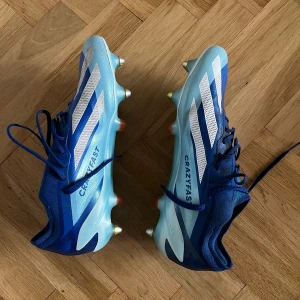 Adidas Crazyfast blå SG - Säljer ett par Adidas Crazyfast fotbollsskor i blått med vita och ljusblå detaljer. Skorna har snörning, spetsig tå och metalldobbar för grymt grepp på planen. Snygg design med klassiska Adidas-ränder och röda loggor på hälen. Perfekta för dig som vill sticka ut på matchen.