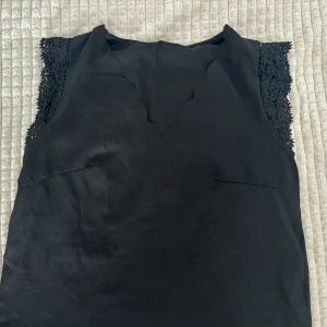Svart  topp med spetsdetaljer - Svart ärmlös topp med snygga spetsdetaljer på axlarna och vågig kant vid halsringningen. Enkel och stilren design som ger en elegant look. Perfekt för dig som gillar klassiska plagg med en twist.