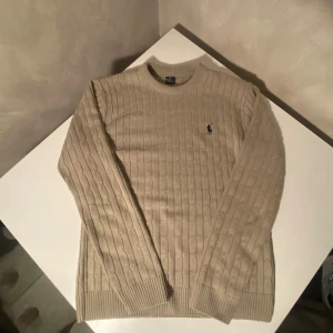 Polo tröja - Kabelstickad beige tröja från Polo Ralph Lauren med klassisk rund halsringning och broderad marinblå logga på bröstet. Tröjan har långa ärmar och ribbade muddar vid ärmslut och nederkant. Perfekt för lager-på-lager under hösten och vintern.
