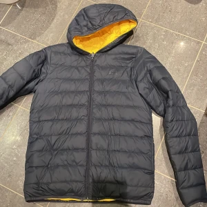 Svart pufferjacka från Hugo Boss - Snygg blå pufferjacka från Hugo Boss med gul insida och huva. Jackan har quiltad design, dragkedja framtill och diskret Boss-logga på bröstet. Perfekt för kalla dagar när du vill ha både stil och värme. Går att vända ut och in. 
