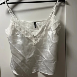 Vit satinlinnetopp med spetsdetaljer - Supersöt vit linnetopp från Gina Tricot i glansigt satinmaterial med tunna axelband och vacker spets längs v-ringningen. Perfekt för att lyfta vilken outfit som helst med en romantisk touch.