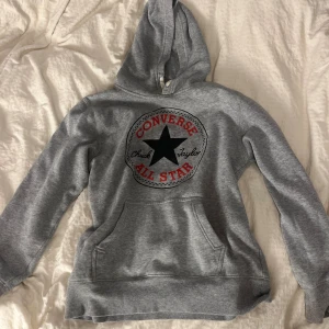 Grå Converse hoodie med logga - Säljer en grå hoodie från Converse med stor klassisk All Star-logga framtill i svart och rött. Tröjan har huva, magficka och ribbade muddar. Perfekt för chill dagar eller när du vill ha en sportig look.