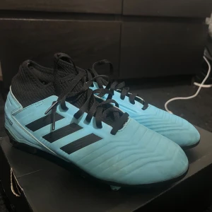 Adidas Predator blå fotbollsskor - Säljer ett par Adidas Predator fotbollsskor i ljusblått med svarta detaljer och snörning. Skorna har en strumpliknande svart ovandel och klassiska tre svarta ränder på sidan. Perfekta för dig som vill sticka ut på planen.