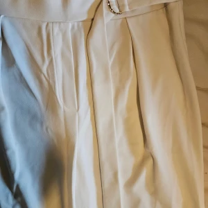 Vita vida byxor med bälte och gulddetalj - Snygga vita byxor med hög midja och ett brett tygbälte med guldfärgad ringdetalj. Byxorna har en rak och lite vidare passform, sydda i ett mjukt och lätt tyg som känns lyxigt. .