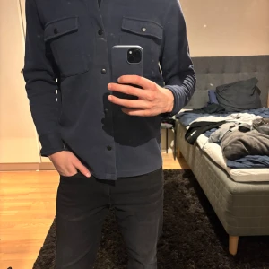 Mörkblå overshirt med knappar - Snygg mörkblå overshirt med två stora bröstfickor och svarta tryckknappar. Skjortan har lång ärm och en clean, stilren look som passar perfekt till jeans. Materialet känns mjukt och bekvämt, och modellen är normal i passformen.