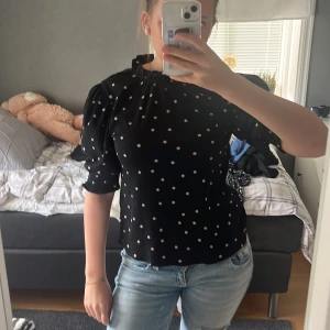 Svart prickig blus med puffärm - Svart blus med vita prickar och puffiga trekvartsärmar. Blusen har en hög, rynkad halsringning och är tillverkad i ett lätt, krinklat tyg som ger en snygg struktur. Perfekt för dig som gillar en söt och trendig look.