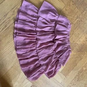 Ljuslila volangkjol med struktur - Supersöt ljuslila kortkjol med volanger och elastisk midja. Kjolen har ett diskret prickigt strukturmönster i tyget och är gjord i ett lätt, luftigt material som känns skönt mot huden. Perfekt för dig som gillar en feminin och trendig stil.