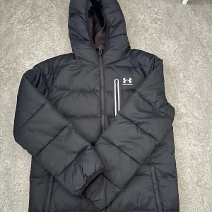 Svart pufferjacka från Under Armour - Svart pufferjacka från Under Armour med huva och halv dragkedja. Jackan har en quiltad design, en bröstficka med dragkedja och logga på vänster sida. Perfekt för kalla dagar och har en sportig vibe. (Pris kan diskuteras vid snabb affär)