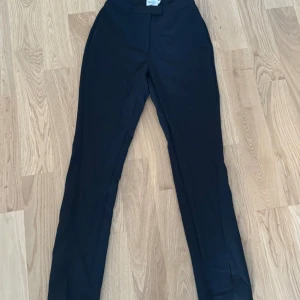 Svarta kostymbyxor från NA-KD med slits - Svarta kostymbyxor från NA-KD med klassisk rak passform och hög midja. Byxorna har dragkedja och knapp framtill samt diskret hake för extra säkerhet. Perfekta för en clean och stilren look.