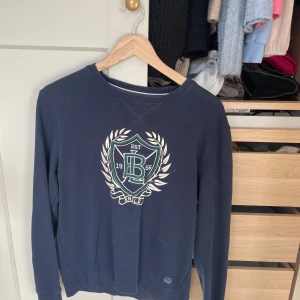 Mörkblå sweatshirt med tryck från Bondelid - Mörkblå sweatshirt från Bondelid med stort tryck framtill i grönt och vitt. Klassisk rund halsringning och långa ärmar. Perfekt för dig som gillar en preppy stil med collegekänsla.