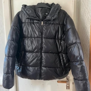 Svart pufferjacka med huva - Säljer en svart pufferjacka från lager 157 med huva och dragkedja framtill. Jackan har en quiltad look med breda kanaler och är perfekt för kyliga dagar. Den har lång ärm och en lite glansig finish som ger en modern vibe.