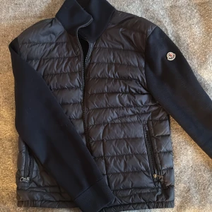 Mörkblå cardigan från Moncler, M - Snygg mörkblå cardigan från Moncler med quiltad framsida och släta ärmar. Jackan har dragkedja framtill, ribbade muddar och Moncler-logga på ärmen. Perfekt för dig som vill ha en stilren och exklusiv look. Den är helt hel och nästan i ny skick!