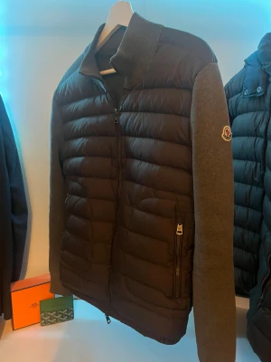 Moncler mörkgrå dunjacka med stickade ärmar - Snygg Moncler dunjacka i mörkgrått med stickade ärmar. Jackan är certifierad enligt DIST och är i nyskick. Klarar sig fint i kalla temperaturer. Priset är självklart inte hugget i sten 