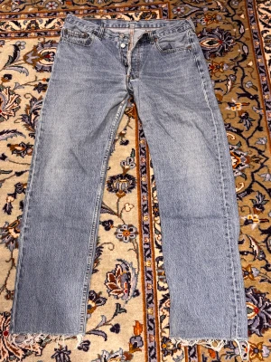 Levi's 501 ljusblå jeans med rå kant - Säljer ett par klassiska Levi's 501 jeans i ljusblå tvätt med raka ben och fransig, rå kant nedtill. Jeansen har fem fickor, knappgylf och den ikoniska läderpatchen bak. Perfekt för dig som gillar vintagevibe och tidlös stil.