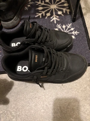 Svarta sneakers från BOSS - Snygga svarta sneakers från BOSS med stilren design och diskreta guldfärgade logodetaljer. Skorna har rund tå, platt sula och snörning. Ovandelen är i slitstark syntet och textil, vilket ger en modern och clean look. Perfekta för dig som gillar minimalistisk stil.