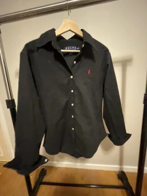 Svart skjorta från Ralph Lauren  - Svart skjorta från Ralph Lauren i storlek XXL. Klassisk modell med lång ärm, vit knappstängning och röd broderad logga på bröstet. Skjortan har en snygg krage och är gjord i mjuk bomull som känns skön mot huden. Perfekt för dig som gillar stilrena plagg med ikonisk detalj.